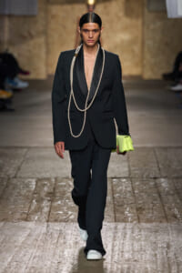 Model walks a runway in a black oversized suit with a deep lapel, layered pearl necklaces, and a bright neon lime clutch, white sneakers visible at the feet