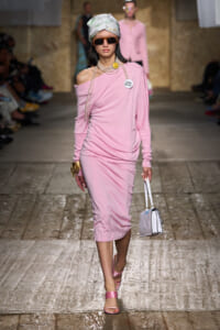 Model walking runway in a pink one-shoulder midi dress with head wrap, sunglasses, and pearl strands, carrying a small white handbag.