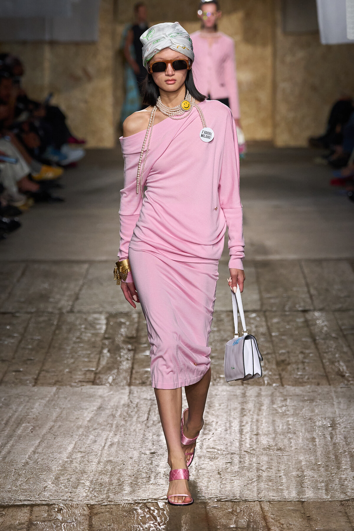 Model walking runway in a pink one-shoulder midi dress with head wrap, sunglasses, and pearl strands, carrying a small white handbag.