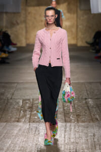 Model walking a runway in a pink cable-knit cardigan over a black midi skirt, accessorized with pearl necklace and bold glasses, carrying a colorful floral bag and wearing rainbow-fluffy shoes.