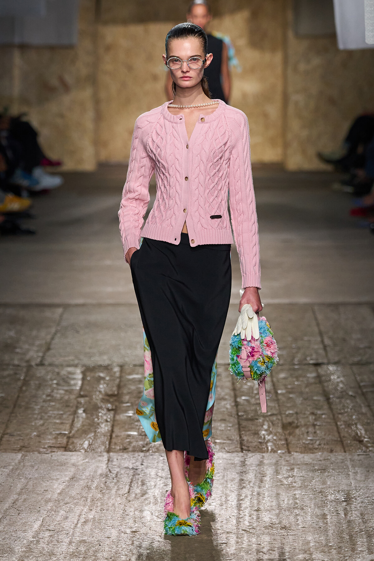 Model walking a runway in a pink cable-knit cardigan over a black midi skirt, accessorized with pearl necklace and bold glasses, carrying a colorful floral bag and wearing rainbow-fluffy shoes.
