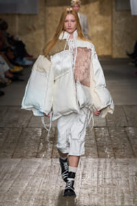 Runway model in an oversized white padded coat with lace, floral panel, and beige accents; black-and-white lace-up boots, industrial backdrop.
