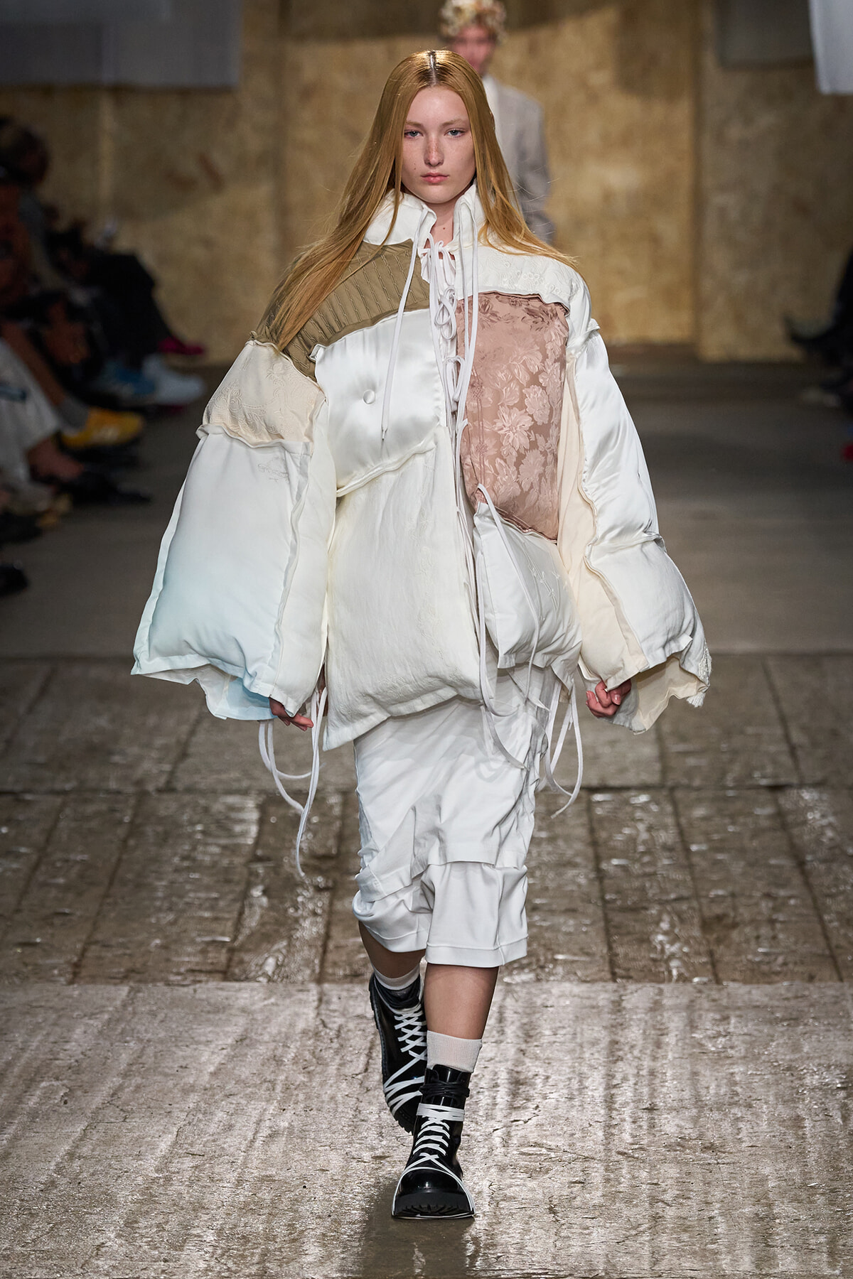 Runway model in an oversized white padded coat with lace, floral panel, and beige accents; black-and-white lace-up boots, industrial backdrop.
