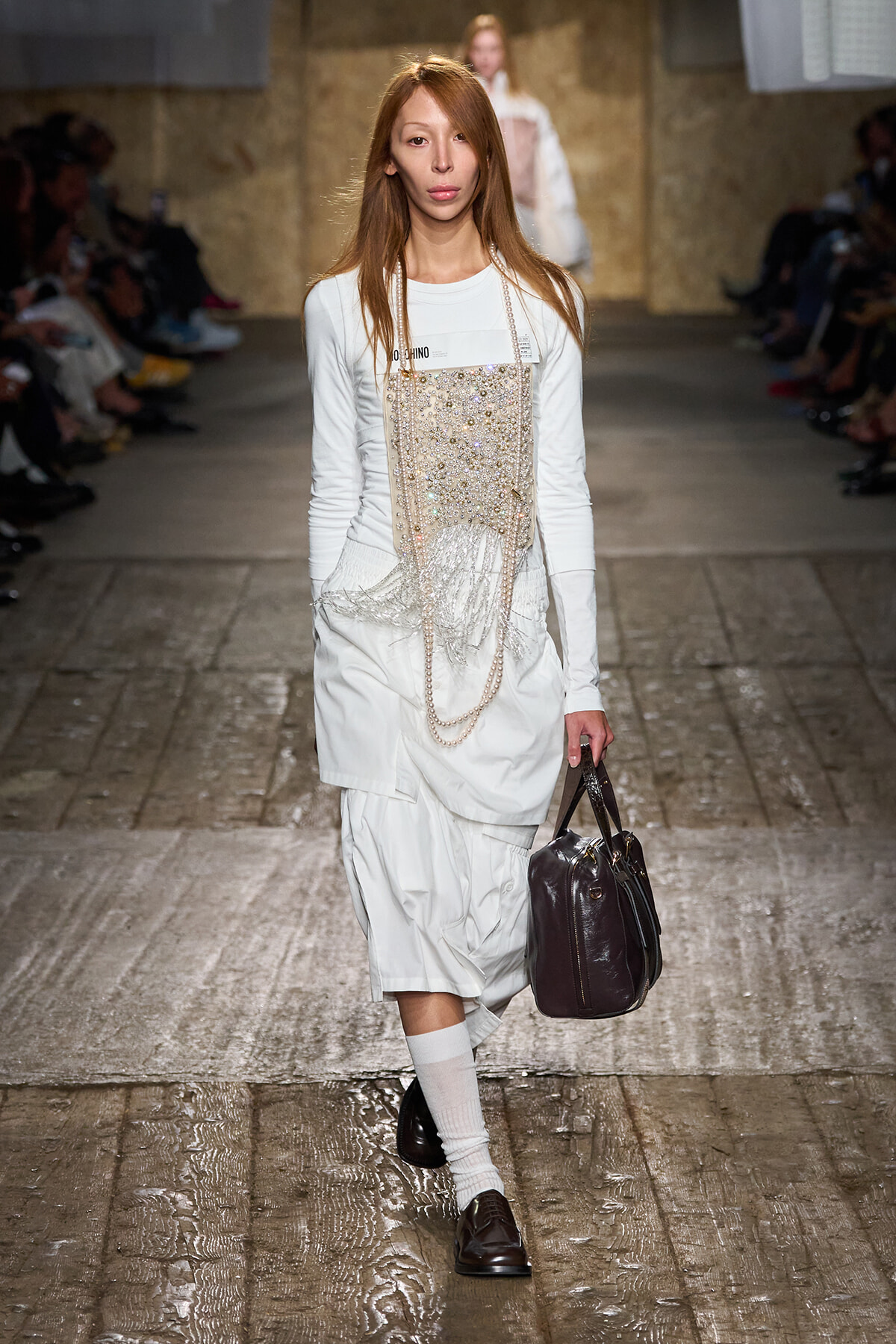 Model walking a fashion show in a white layered outfit with beaded fringe and pearl chains, carrying a dark brown handbag.