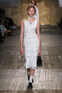 Model walking a white, sculpted dress with string wrap details on a runway, carrying a small black bag.