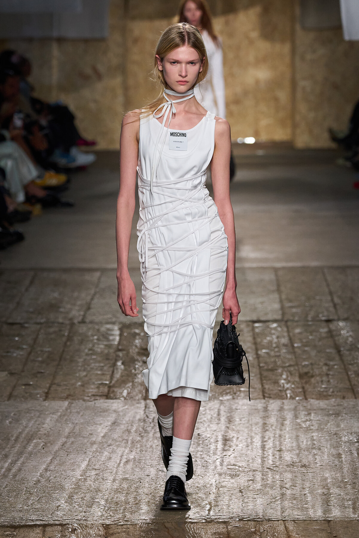 Model walking a white, sculpted dress with string wrap details on a runway, carrying a small black bag.