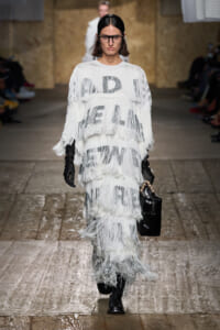 Runway model in a white, multi-layer fringe dress with bold black lettering, carrying a black handbag and wearing black gloves.