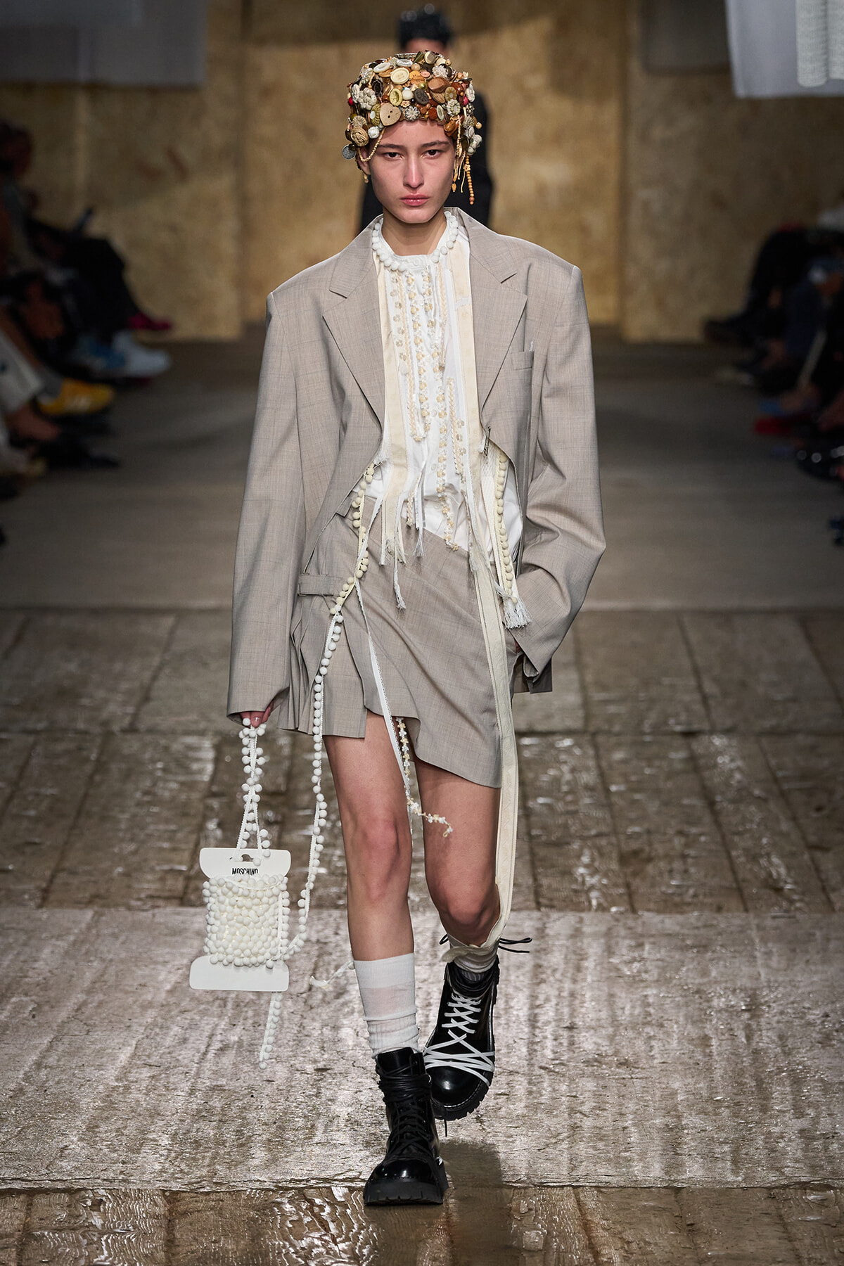 Model on a runway wearing a light beige suit with a cream layered blouse, shell headpiece, and pearl-bead handbag, paired with black combat boots.