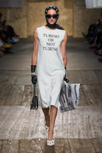 Model walks the runway in a sleeveless white dress bearing the text 'TUBINO OR NOT TUBINO,' with a black polka-dot headscarf, oversized sunglasses, black gloves, and pearl bracelets, carrying a small handbag.