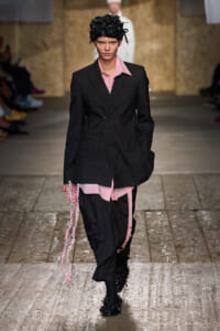 Model walking a runway in a menswear-inspired black pinstripe suit with pink shirt accents and a sculpted black headpiece in a dimly lit venue.