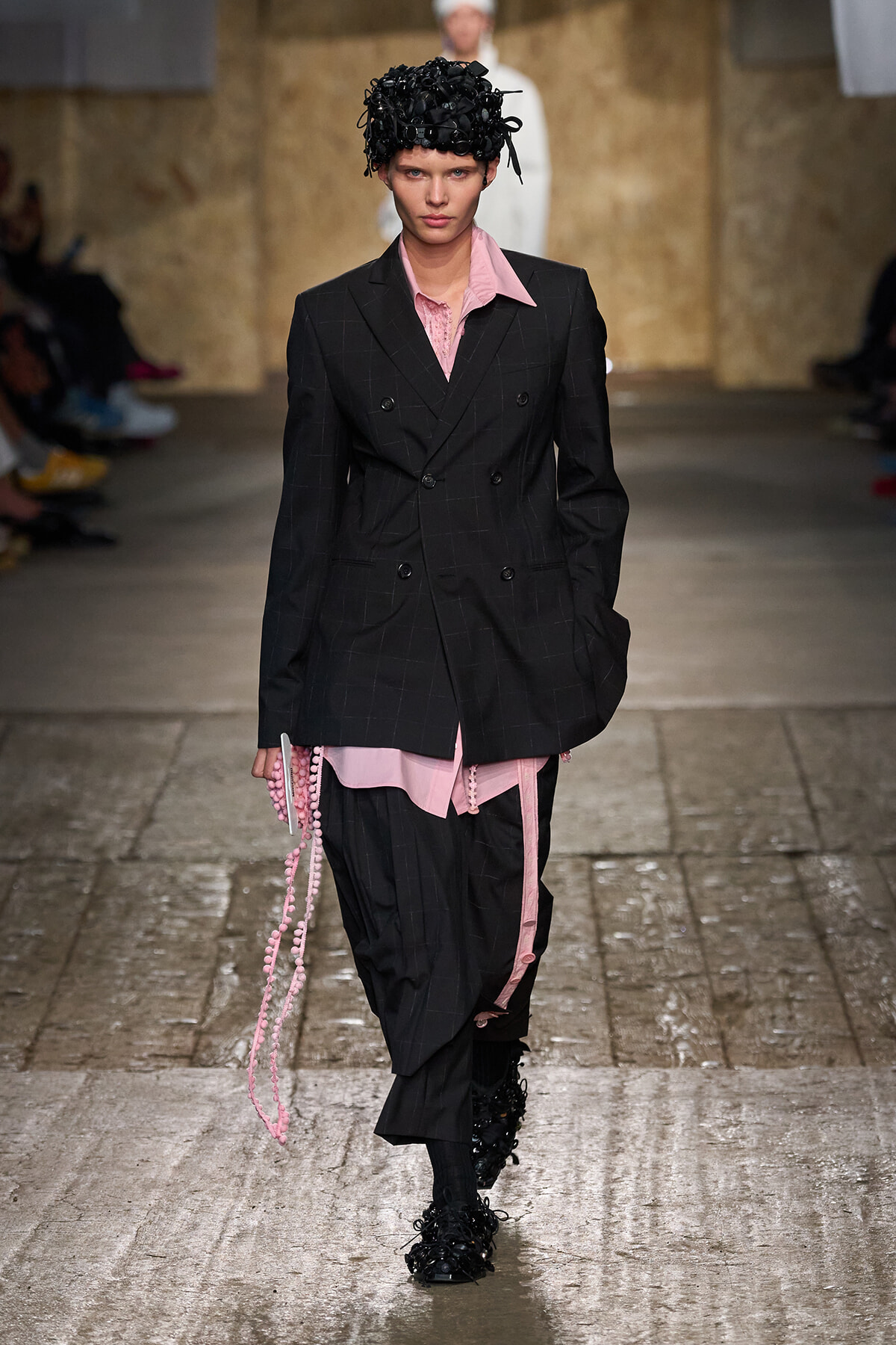 Model walking a runway in a menswear-inspired black pinstripe suit with pink shirt accents and a sculpted black headpiece in a dimly lit venue.