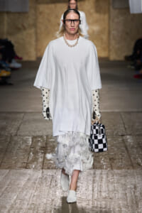 Model walks the runway in an oversized white poncho dress with a feathered fringe skirt, pearl necklace, polka-dot gloves, and a black‑and‑white checkered bag.