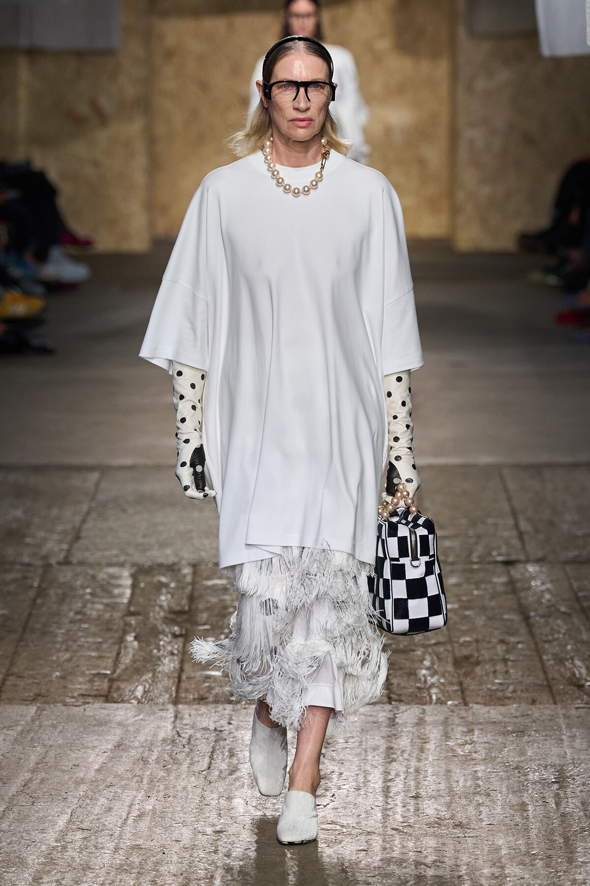 Model walks the runway in an oversized white poncho dress with a feathered fringe skirt, pearl necklace, polka-dot gloves, and a black‑and‑white checkered bag.