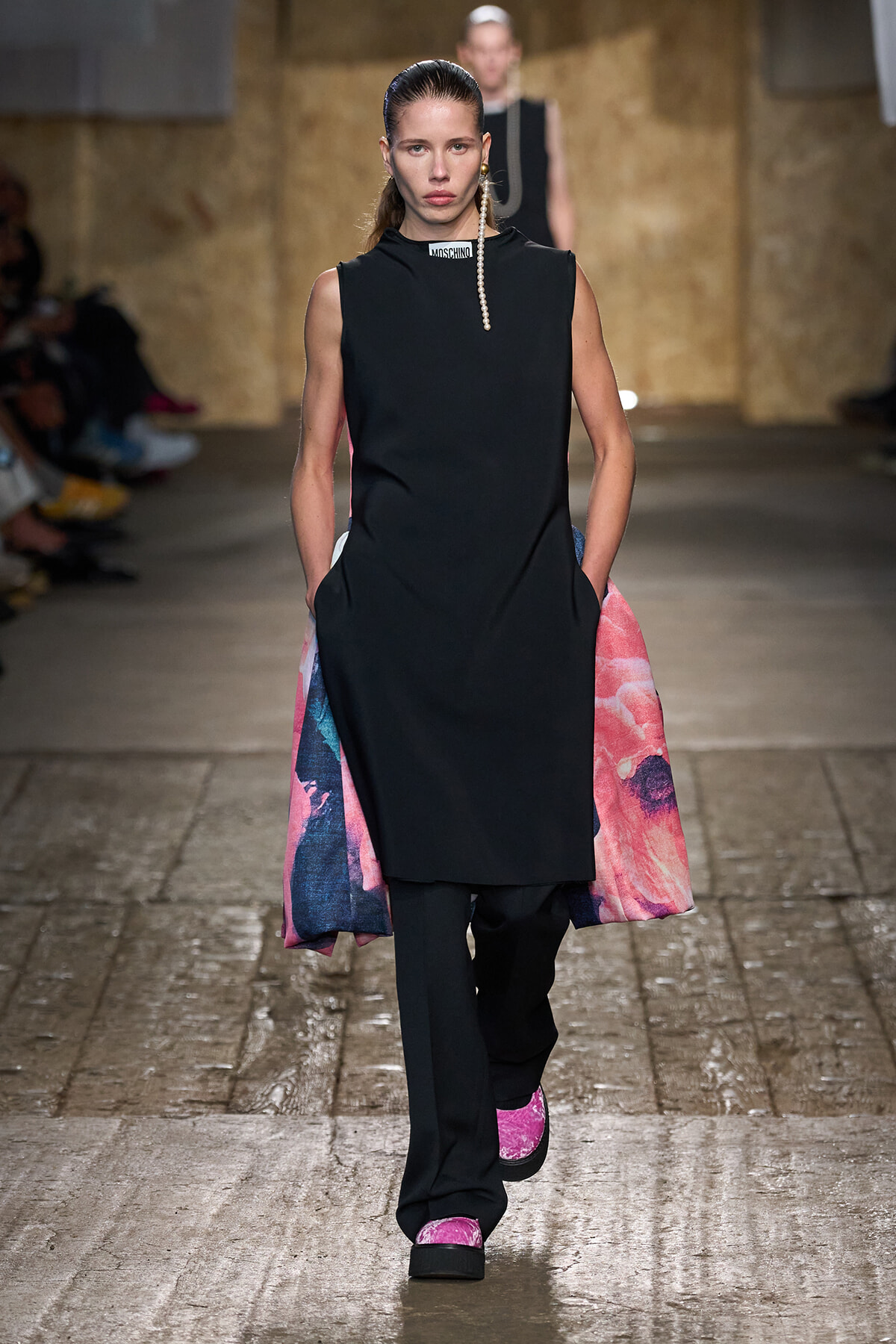 Model walking a runway in a sleeveless black tunic over black trousers, with bright pink‑blue side panels and pink platform shoes, pearl earring visible.