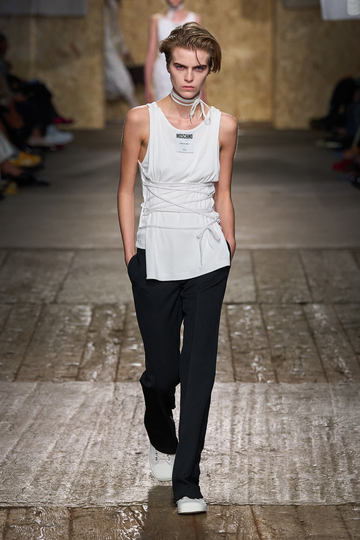 Model walks a fashion runway in a white sleeveless top with wrap-around laces over black pants and white sneakers.