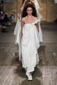 Model strolling a runway in a white, off-the-shoulder gown with a knot at the bust, hands in pockets, embroidered hem, and white loafers.