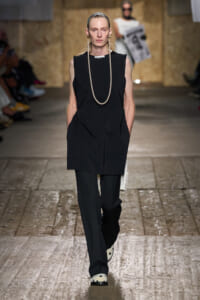 Model walking a fashion runway in a black sleeveless tunic over black trousers, accessorized with a long pearl necklace and white polka-dot shoes.