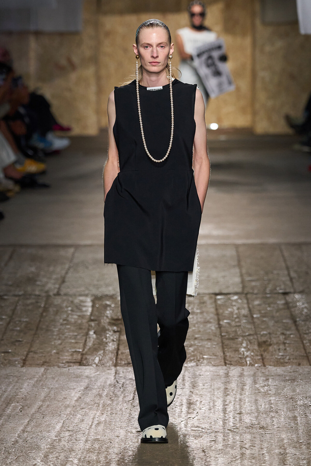 Model walking a fashion runway in a black sleeveless tunic over black trousers, accessorized with a long pearl necklace and white polka-dot shoes.
