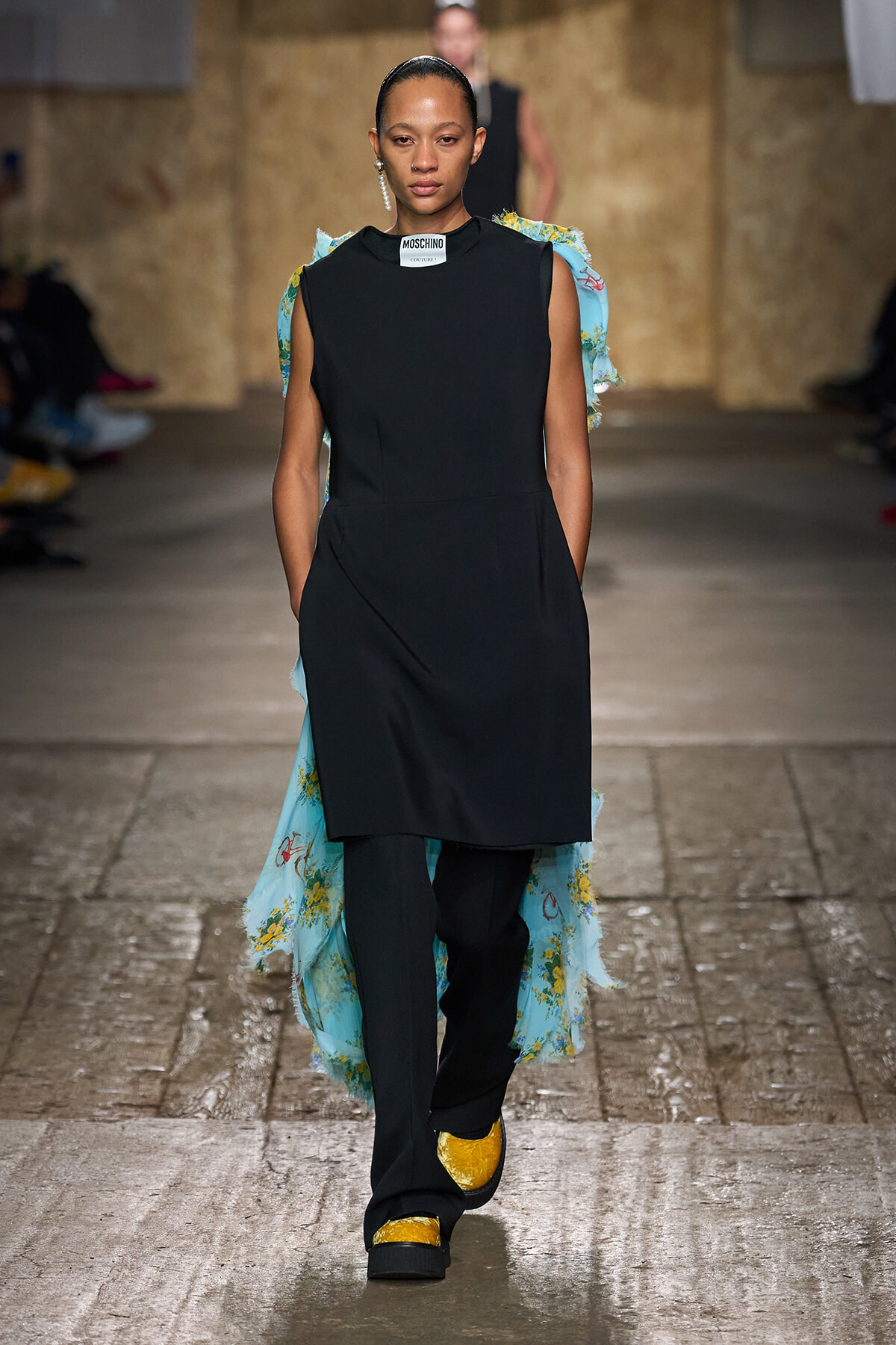 Female model walking a runway in a black sleeveless tunic over pants, with a light blue floral cape and yellow shoes.