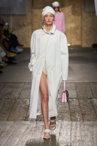Model walks the runway in a long white coat, white head wrap, and beige sandals, carrying a pink handbag.