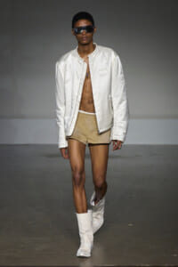 Model on a runway wearing an oversized white bomber jacket, tan shorts, white boots, and large dark sunglasses.