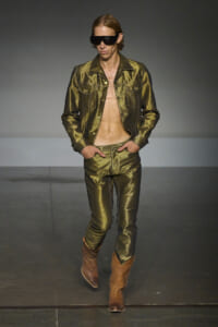 Male model walks a fashion runway in a metallic olive-green denim jacket and matching pants, with an open chest, brown boots, and oversized sunglasses.