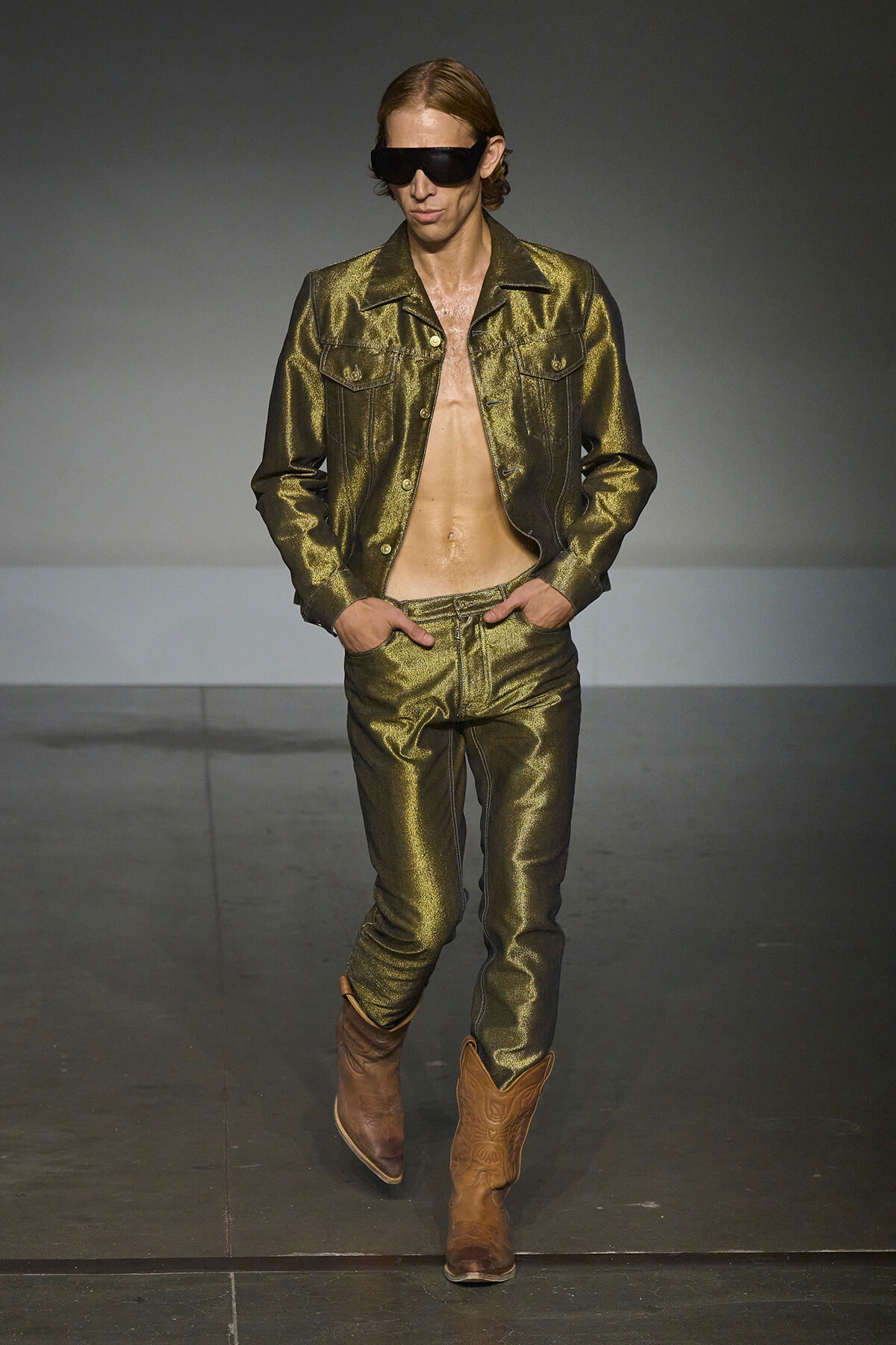 Male model walks a fashion runway in a metallic olive-green denim jacket and matching pants, with an open chest, brown boots, and oversized sunglasses.