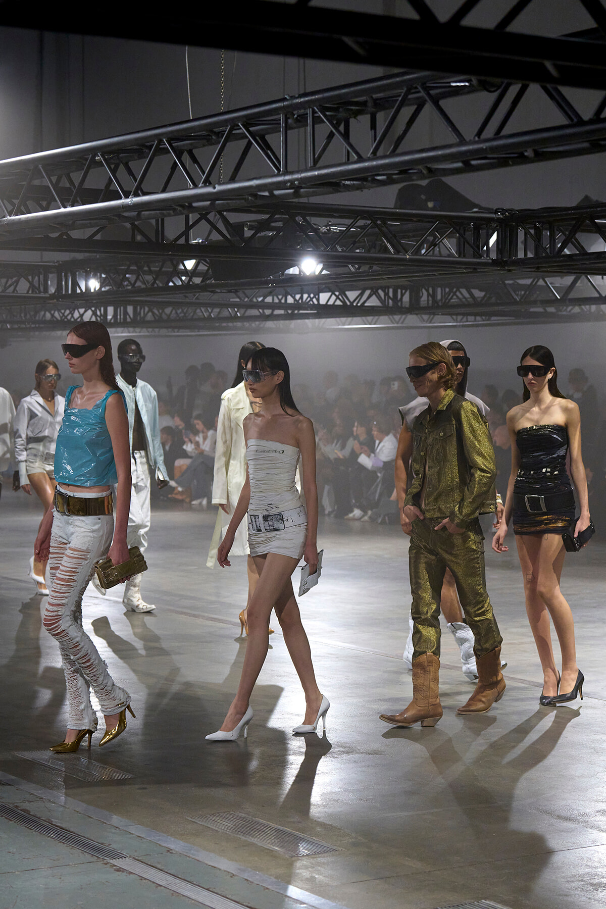 Models walk a fashion show runway in metallic and white outfits with futuristic sunglasses, under exposed truss lighting.