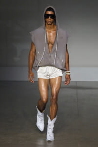 Male model on a runway wearing a gray sleeveless hooded top with zip, white shorts, and white boots, plus black sunglasses.