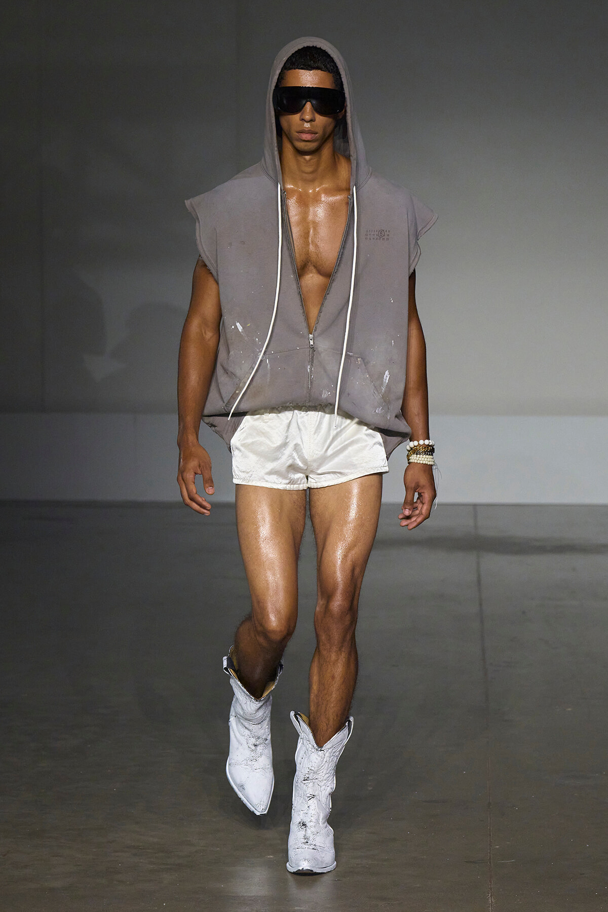 Male model on a runway wearing a gray sleeveless hooded top with zip, white shorts, and white boots, plus black sunglasses.
