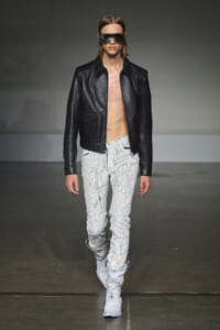 Male model on a runway wearing a black leather jacket, no shirt, white splattered jeans, and white boots with futuristic sunglasses