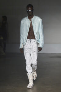 Male model on a runway wearing a pale blue bomber jacket, white paint-splattered jeans, and chunky white boots with oversized sunglasses.