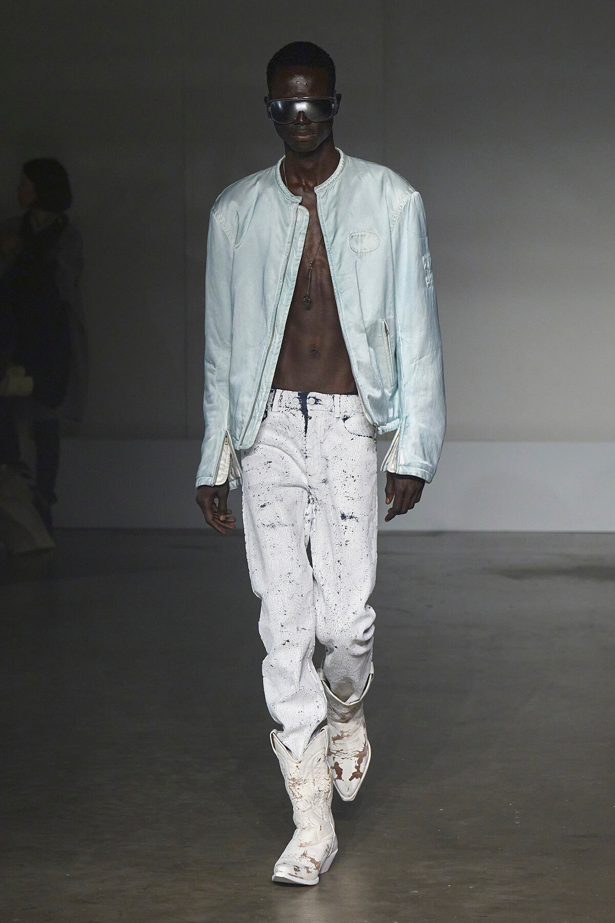 Male model on a runway wearing a pale blue bomber jacket, white paint-splattered jeans, and chunky white boots with oversized sunglasses.