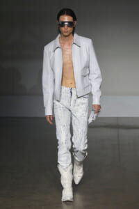 Male model on a runway wearing a light gray zip jacket, splattered pale jeans, white boots, and futuristic sunglasses on a dark backdrop.
