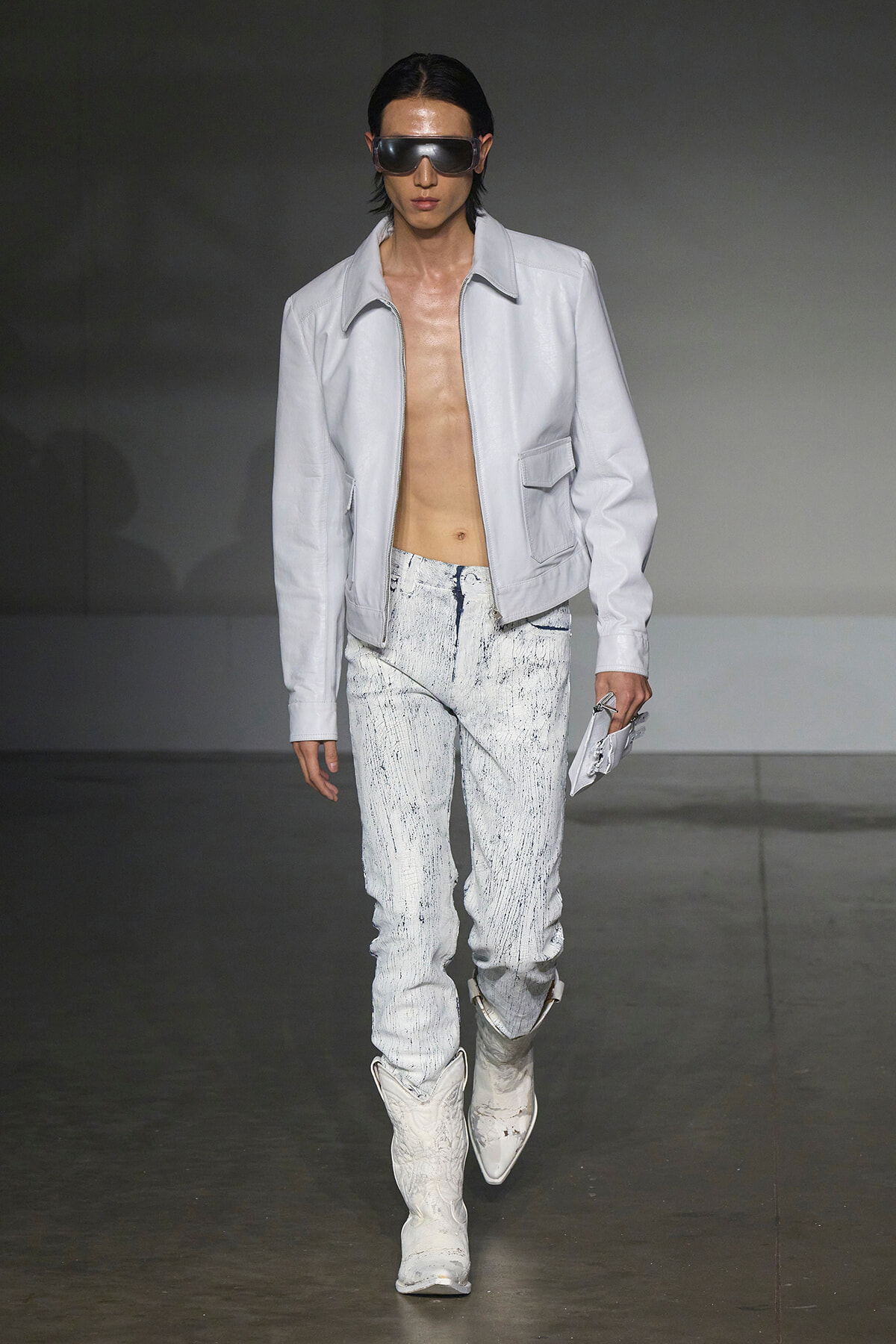 Male model on a runway wearing a light gray zip jacket, splattered pale jeans, white boots, and futuristic sunglasses on a dark backdrop.