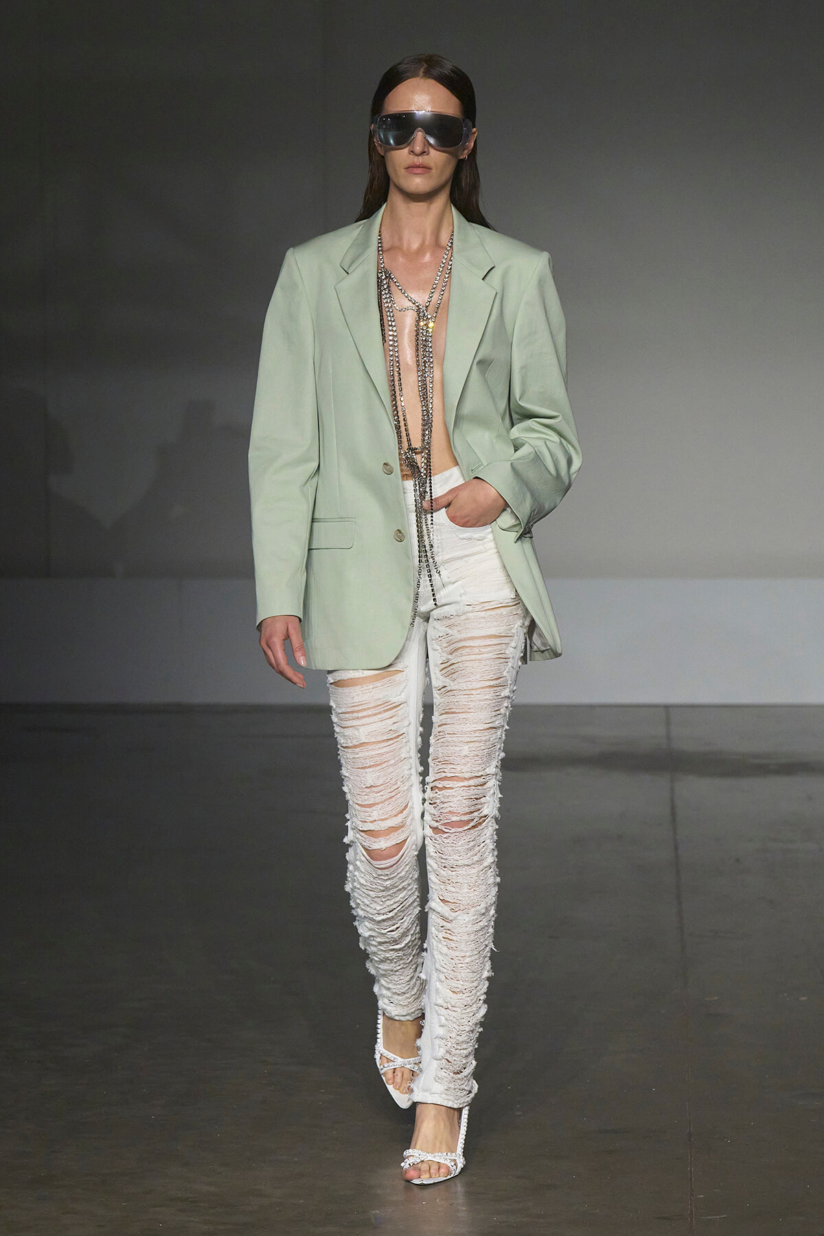 Model on runway wearing a mint oversized blazer, white ripped jeans, layered chains, and oversized sunglasses.