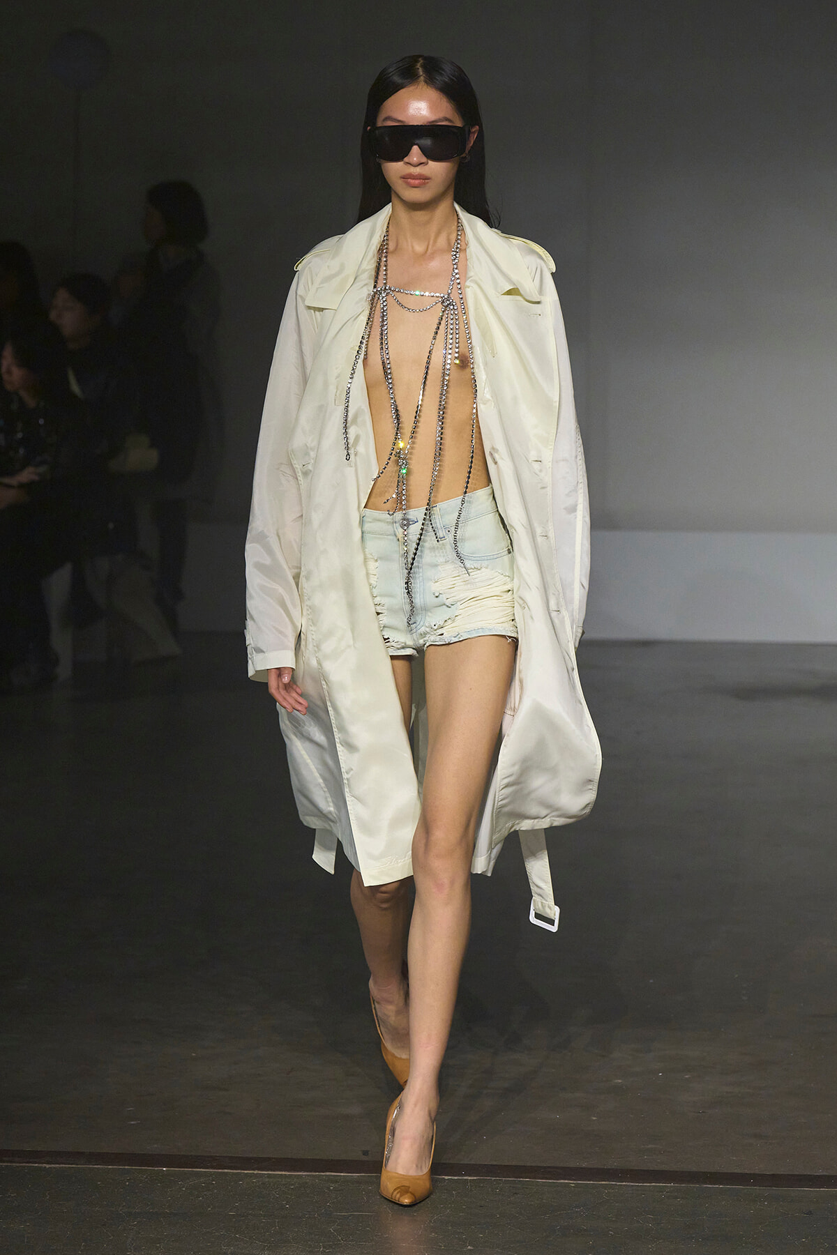 Model on a runway wearing a long pale trench coat, oversized sunglasses, and layered silver chains over distressed denim shorts with tan heels.
