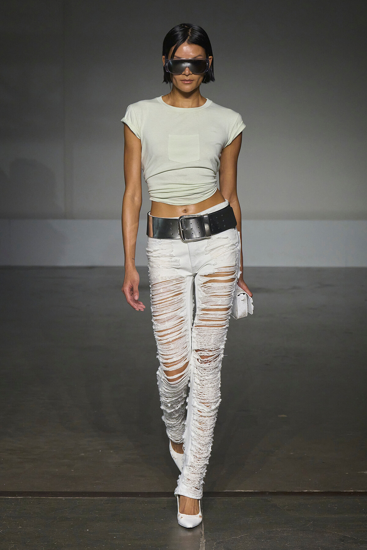 Model walking a runway in a pale cropped tee, ultra-ripped white jeans, wide black belt, white heels, and oversized sunglasses.