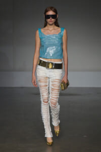 Model on a runway wearing a metallic belt, light blue plastic crop top, and heavily distressed white jeans with gold flats, black sunglasses, and a gold clutch.