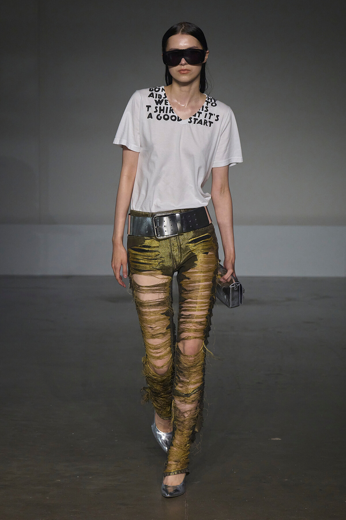 Runway model in a white graphic tee, oversized black belt, gold shredded pants, silver heels, and large sunglasses with a small clutch.