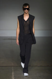 Male model walks the runway in a black sleeveless blazer over a sheer black top, black trousers, white sneakers, and oversized sunglasses.