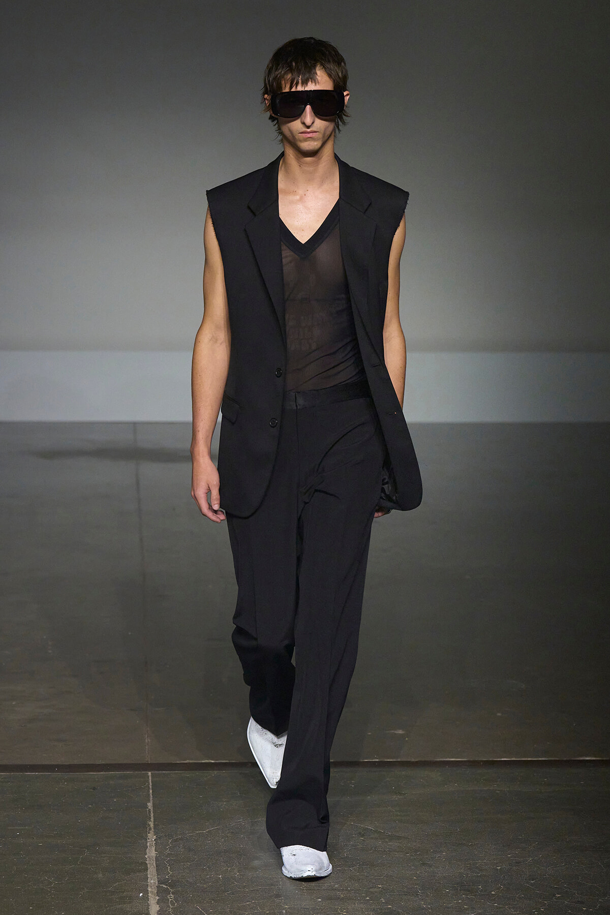 Male model walks the runway in a black sleeveless blazer over a sheer black top, black trousers, white sneakers, and oversized sunglasses.