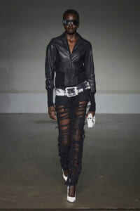 Model walking a fashion show in a black leather jacket, oversized white belt, shredded black pants, white heels, and sunglasses with a white clutch accessory.