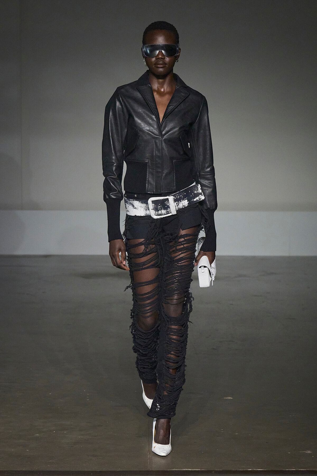 Model walking a fashion show in a black leather jacket, oversized white belt, shredded black pants, white heels, and sunglasses with a white clutch accessory.
