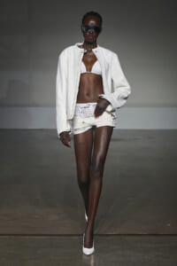 Model walking runway in a white jacket, white bikini top, and distressed white shorts with a wide belt and white heels, wearing oversized sunglasses.