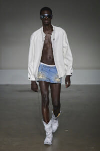 Male model walking a runway in a white jacket, ripped denim shorts, and white boots with dark sunglasses.