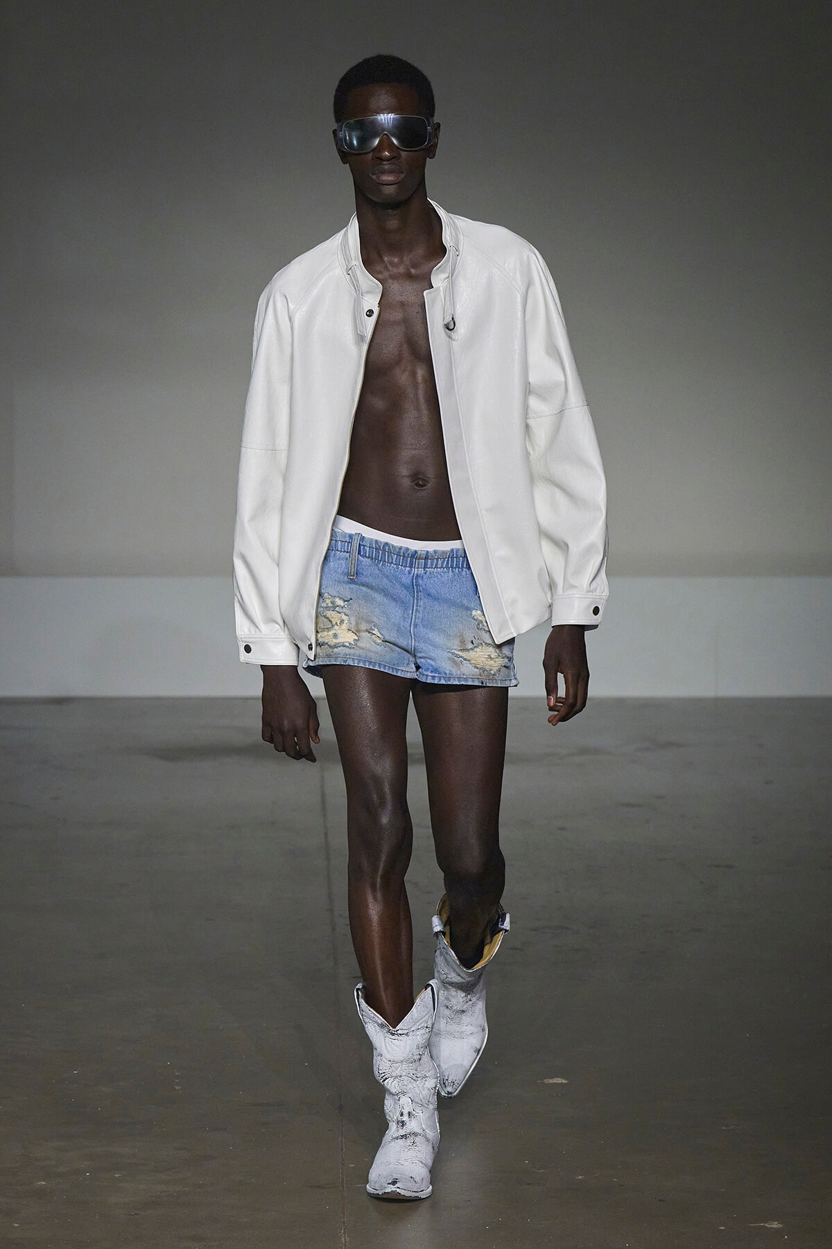 Male model walking a runway in a white jacket, ripped denim shorts, and white boots with dark sunglasses.