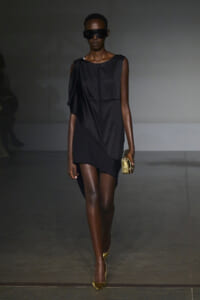 Model on a runway wearing a sleeveless black asymmetrical dress, oversized black sunglasses, and gold clutch with matching gold heels against a gray backdrop.