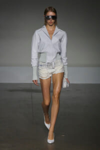 Model walking a runway in a light gray utility jacket, white distressed shorts, wide belt, oversized sunglasses, and white heels.