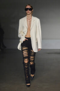 Model walking a runway in an oversized cream blazer, extremely ripped black jeans, black pointed heels, and oversized dark sunglasses.